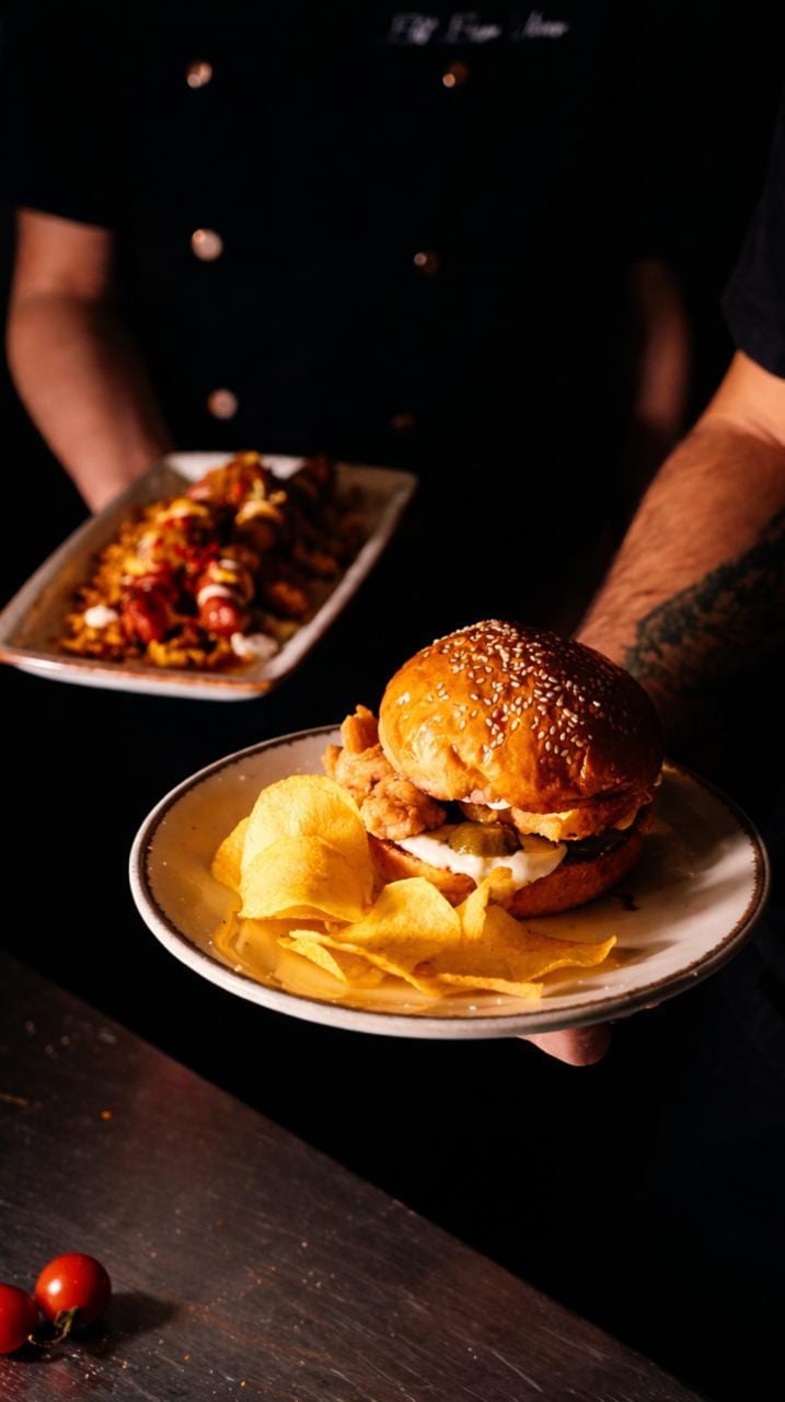 Garsonun elinde susamlı burger ve cips, arkada başka bir tabakta soslu atıştırmalık servis anı