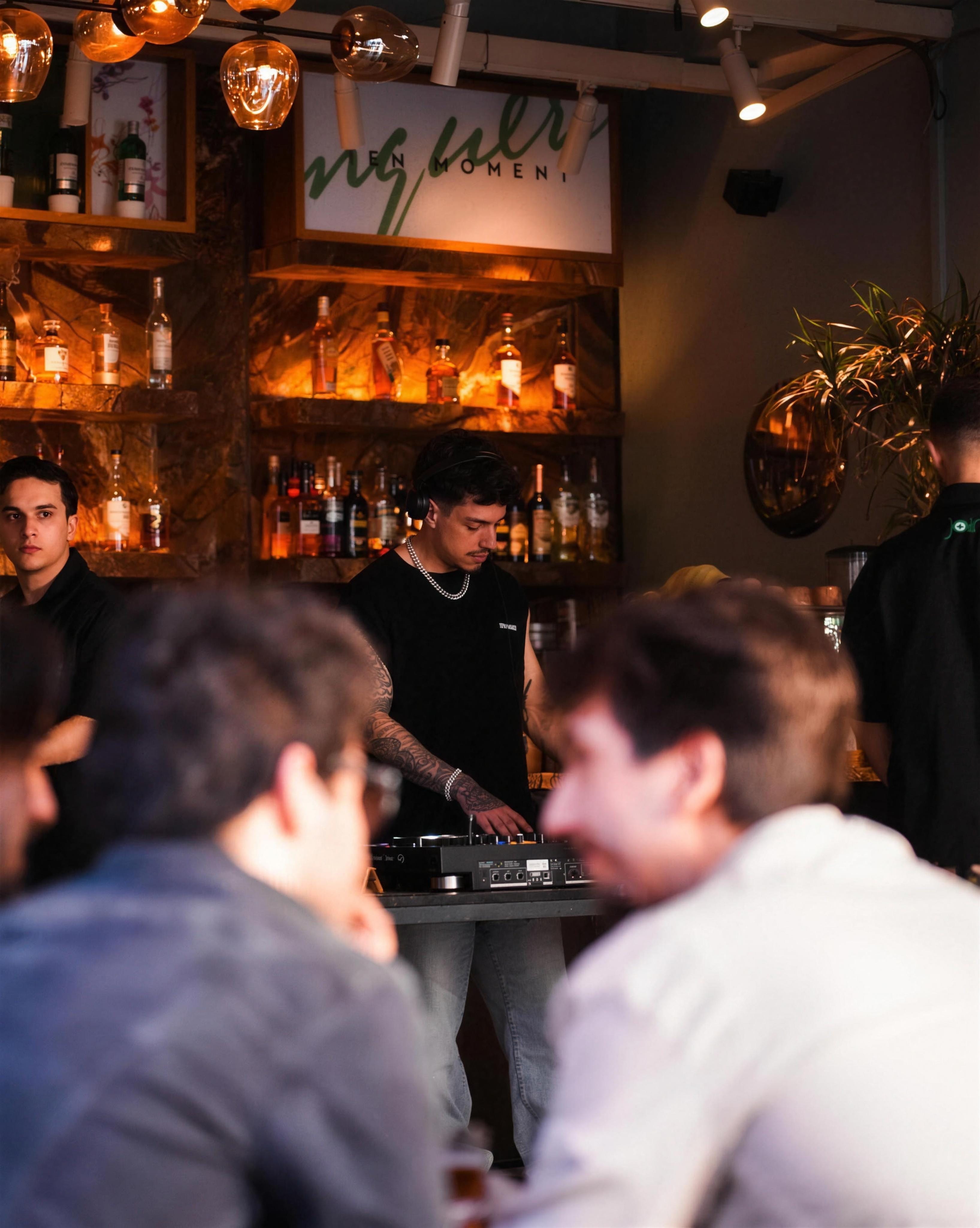 Gece mekan içi, bar arkasında DJ setinde performans veren DJ ve önde oturan misafirler, sıcak ışıklı şişe rafları