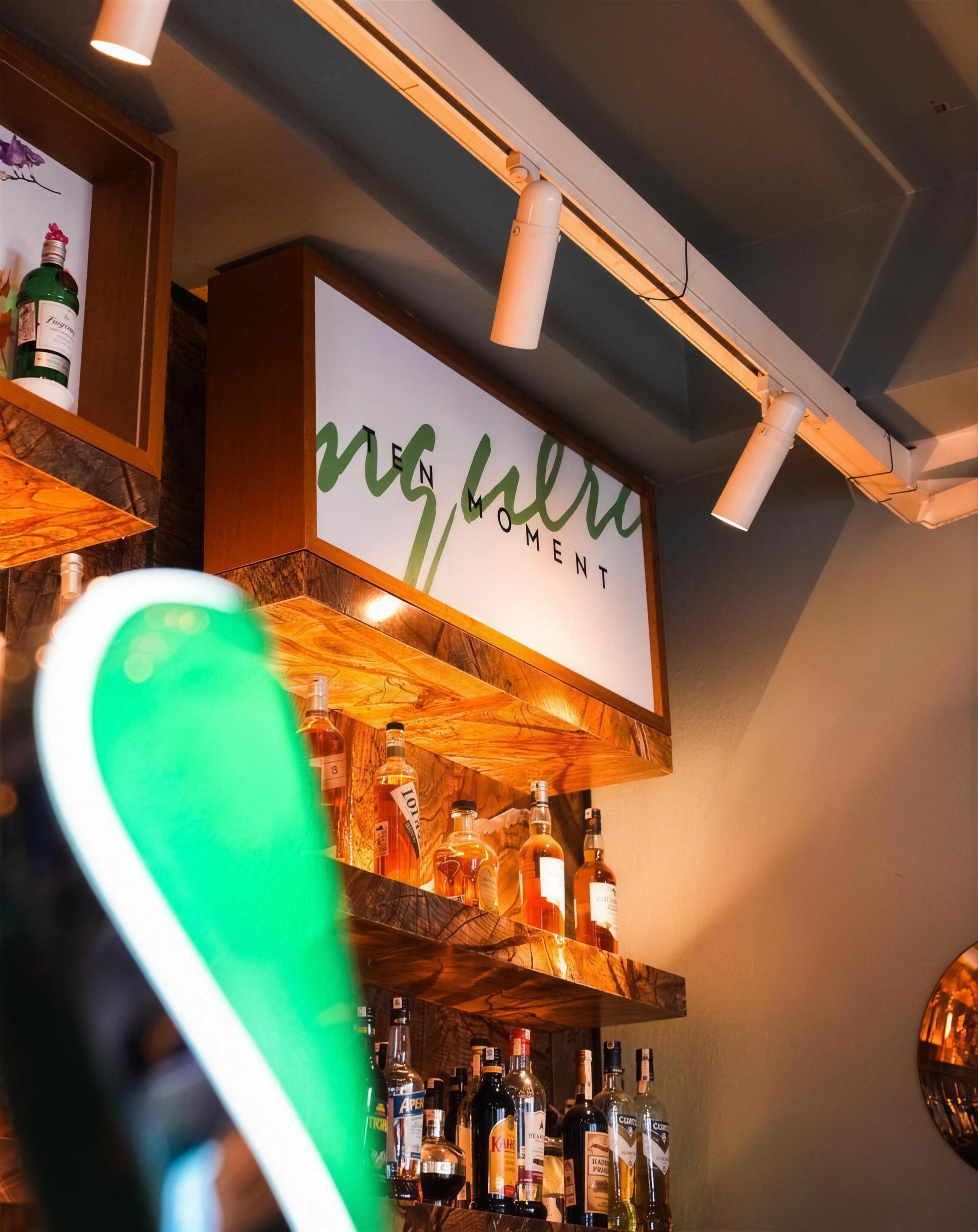 Bar rafları üzerinde asılı ışıklı tabela, ön planda yeşil neon ışık bulanık, arka planda şişe rafları